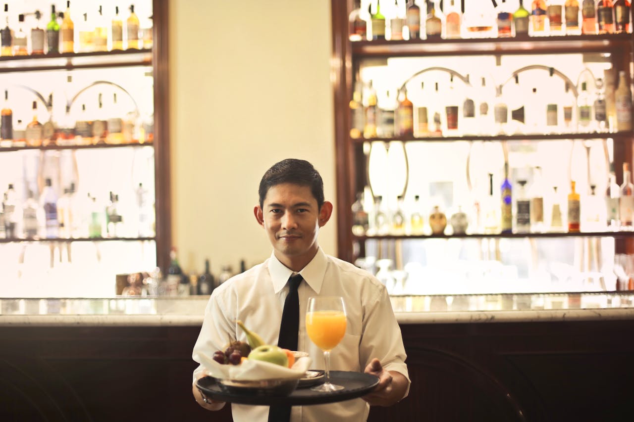 Waiter serving a drink and fruit tray in a bar with bottles displayed in the background.